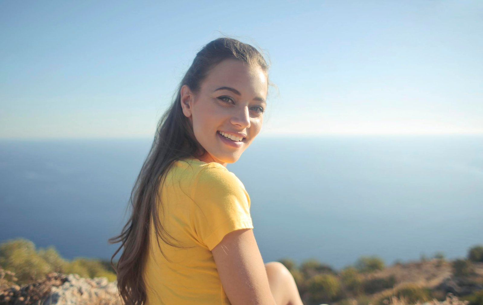 Young woman in a yellow shirt smiles by the sea, enjoying the outdoor view.